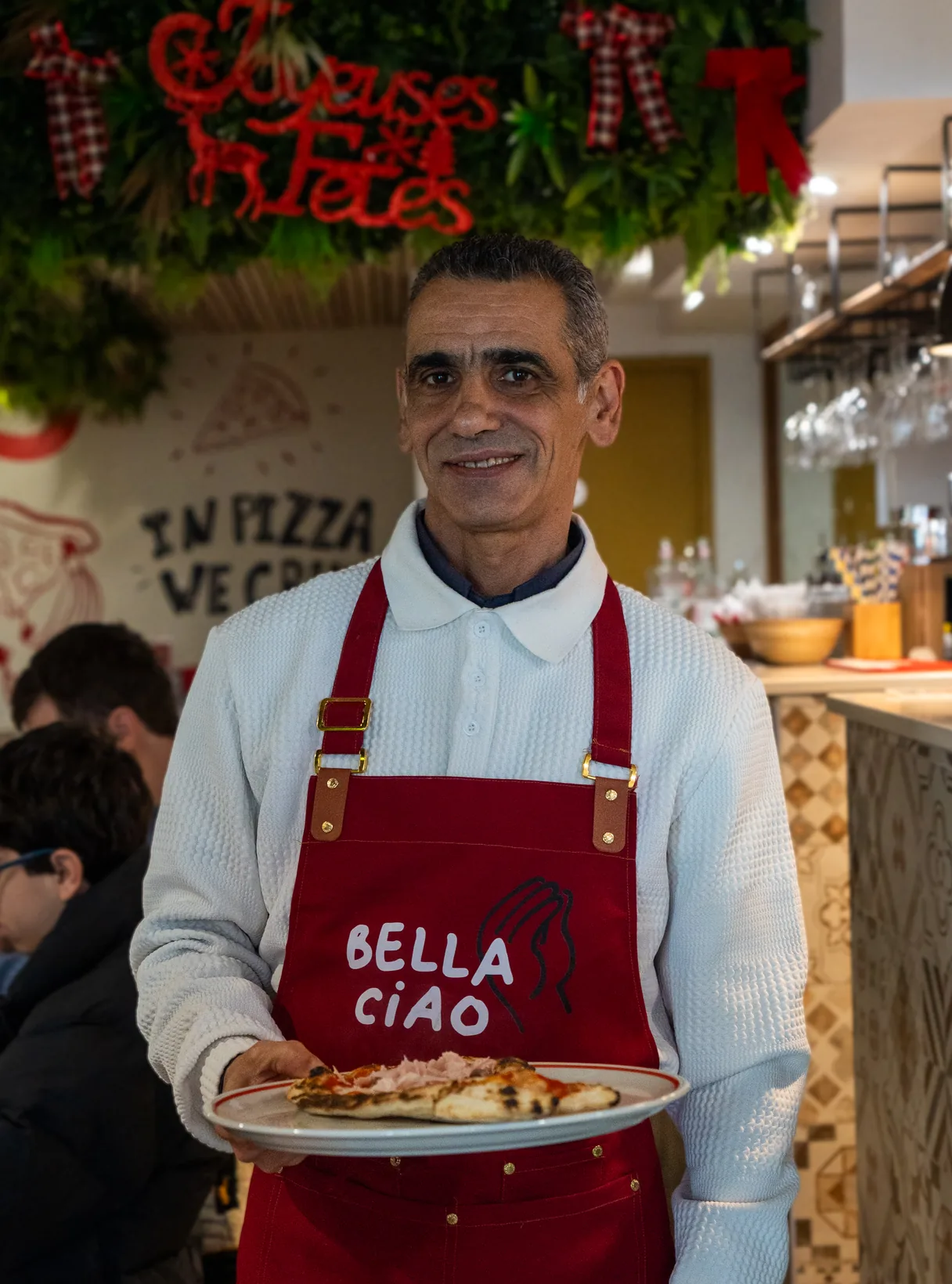 A Bella Ciao waiter in the red logo apron serving a pizza, the In pizza we crust wall behind him