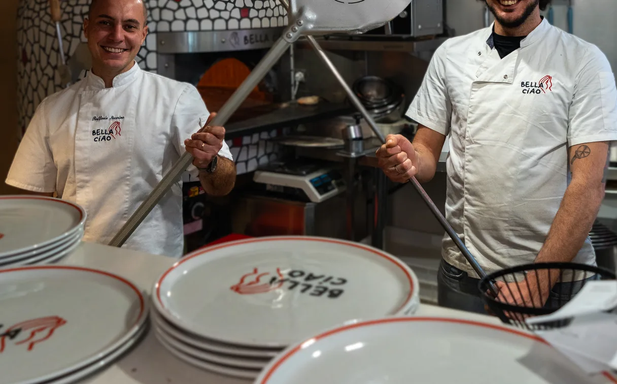 Two pizzaioli at the oven with a Bella Ciao peel, the plates printed with the logo in front of them